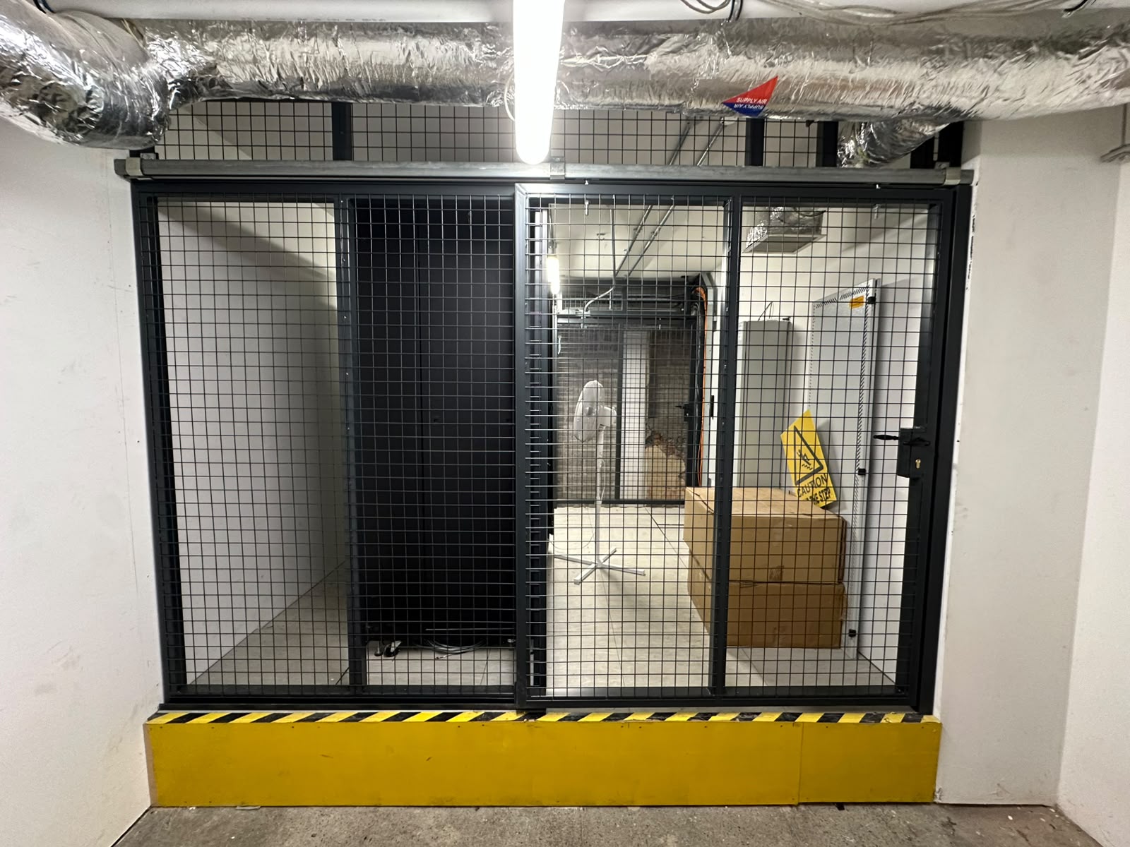 Dark grey welded mesh security cage with sliding gate enclosing a plant room, with yellow kerb protection at floor level